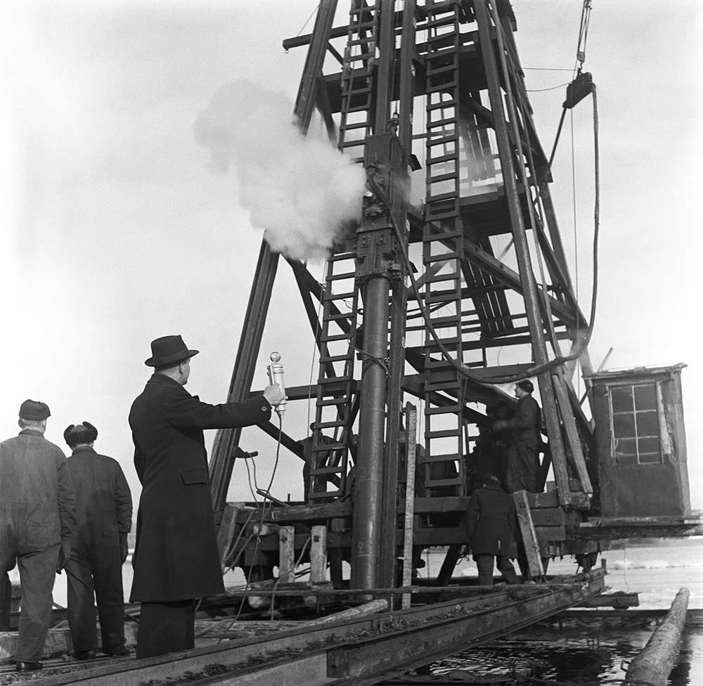 A radio sound recordist in a long coat and hat holds a microphone up to a steaming pile driver on a frozen harbour, 1930s