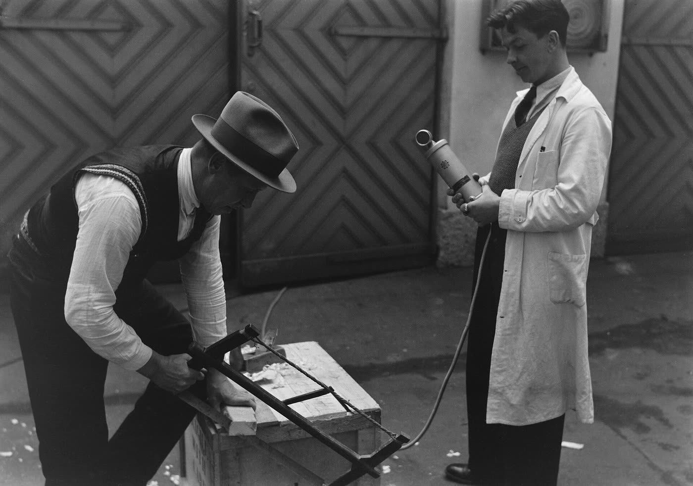Two men in a yard in front of diamond-patterned doors: one saws a plank on a wooden crate while the other, in a white coat, holds a microphone towards the saw